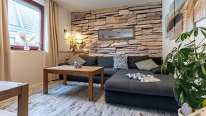 A cozy living room with a stone wall, a dark grey sofa, a wooden coffee table, and a large window with curtains. - Ferienwohnung in Busum (Büsum)