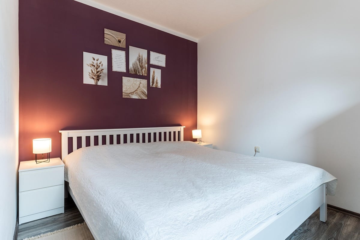 A bedroom with a white bed, a nightstand with a lamp, and a wall with framed pictures.
