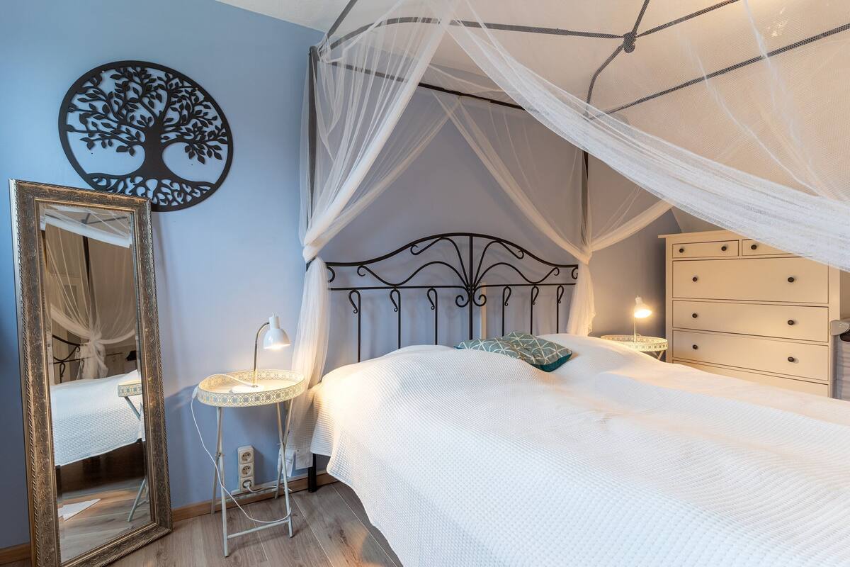 A bedroom with a canopy bed, a bedside table with a lamp, a dresser, and a wall art piece.