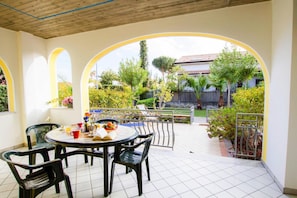 A covered patio with a round table and chairs, surrounded by arches, and a view of a garden and buildings in the background. - Apartment in Cilento Near Sandy Beach (Ascea)