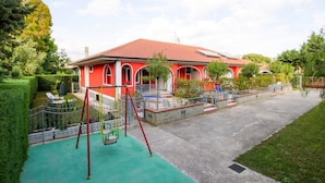 Una casa roja con un patio de juegos verde al frente, rodeada de árboles y un cielo despejado.