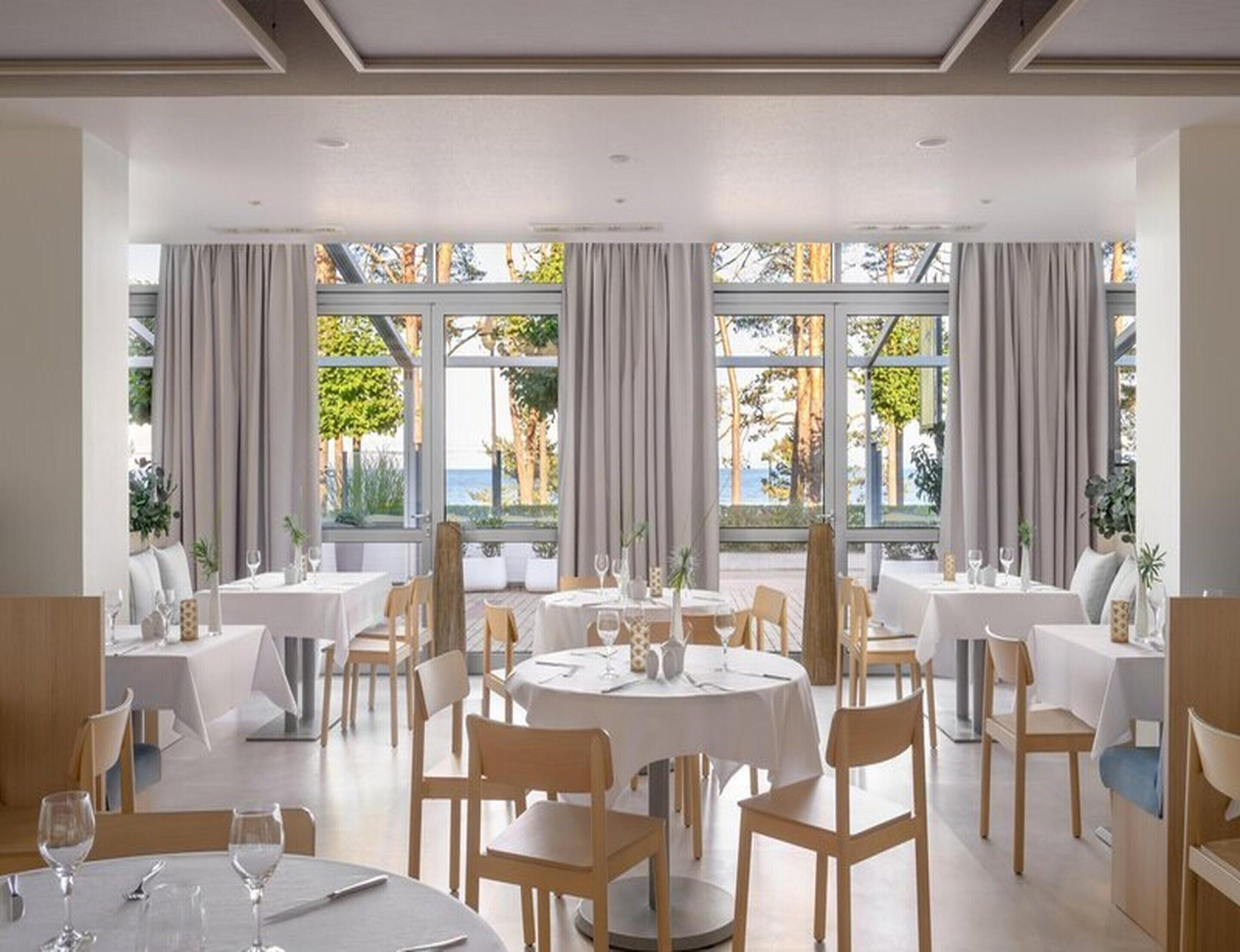 A restaurant with round tables and chairs, large windows with curtains, and a view of the outdoors.