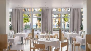 A restaurant with round tables and chairs, large windows with curtains, and a view of the outdoors.