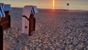 A beach with two striped beach chairs and a sunken chair, a clear view of the sunset over the water, and a sign in the distance. - Appartements im Ferienpark Rugen in Binz (Binz)