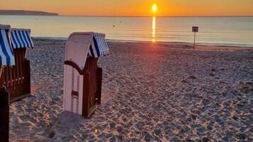 Una playa con dos sillas de playa a rayas y un sillón hundido, una vista despejada del atardecer sobre el agua, y un cartel a lo lejos.