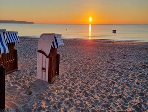 En strand med to stribede liggestole og en nedsænket stol, et klart syn af solnedgangen over vandet, og et skilt i det fjerne.
