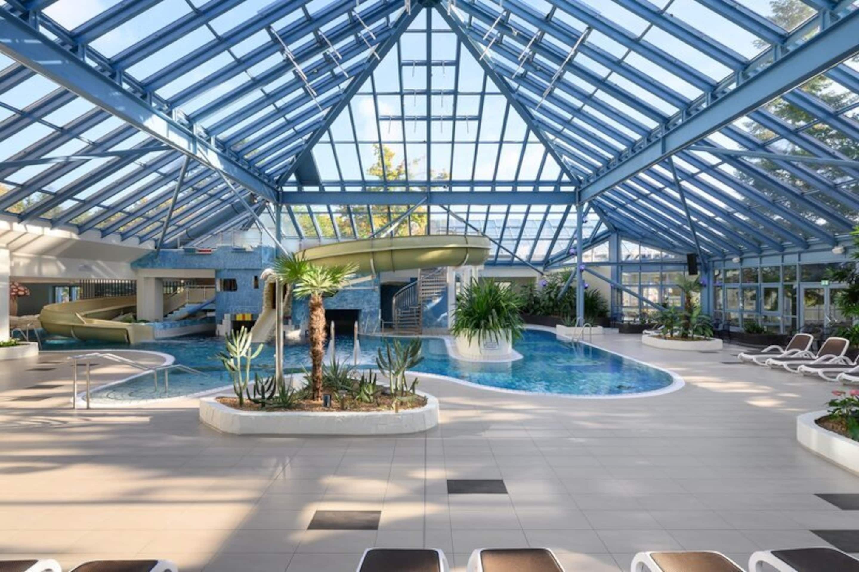 En indendørs swimmingpool med glasloft, liggestole og en vandrutsjebane.