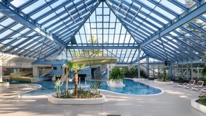 An indoor swimming pool with a glass ceiling, lounge chairs, and a water slide. - Appartements im Ferienpark Rugen in Binz (Binz)