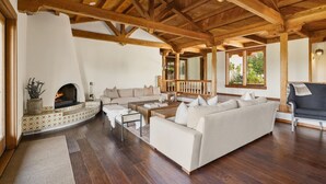 A spacious living room with a fireplace, wooden beams, a sofa, and a view of greenery outside.