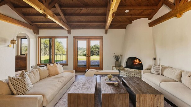A cozy living room with a fireplace, wooden beams, and a view through large windows.