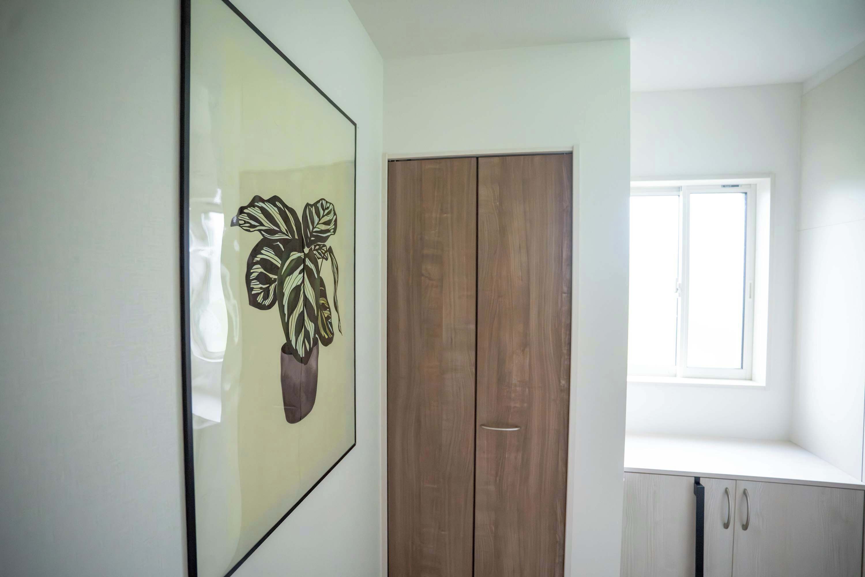 A room with a wooden wardrobe, a framed artwork on the wall, and a window providing natural light.