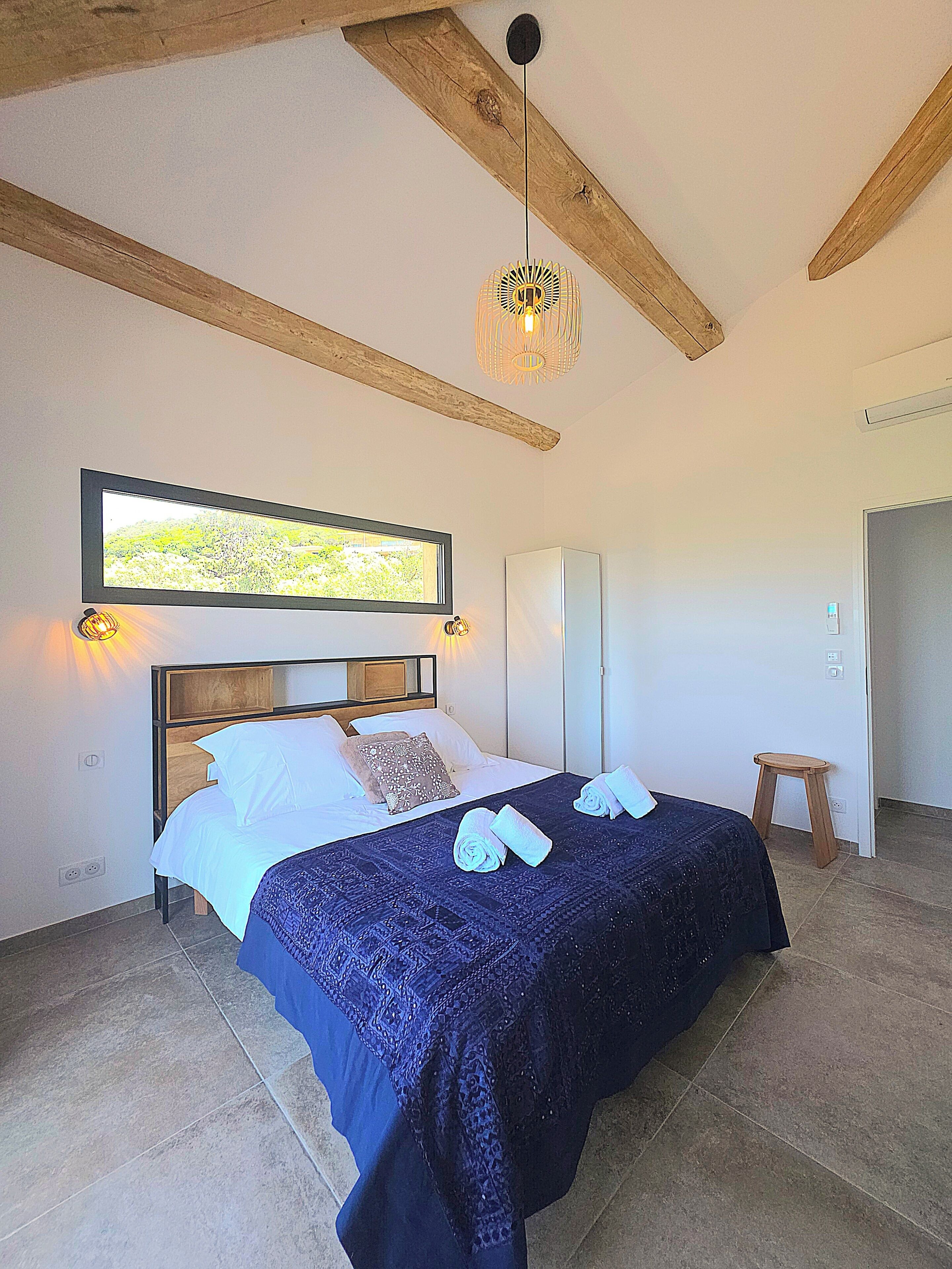 Un dormitorio con cama, viga de techo de madera, lámpara colgante, un cuadro en la pared y un taburete pequeño.