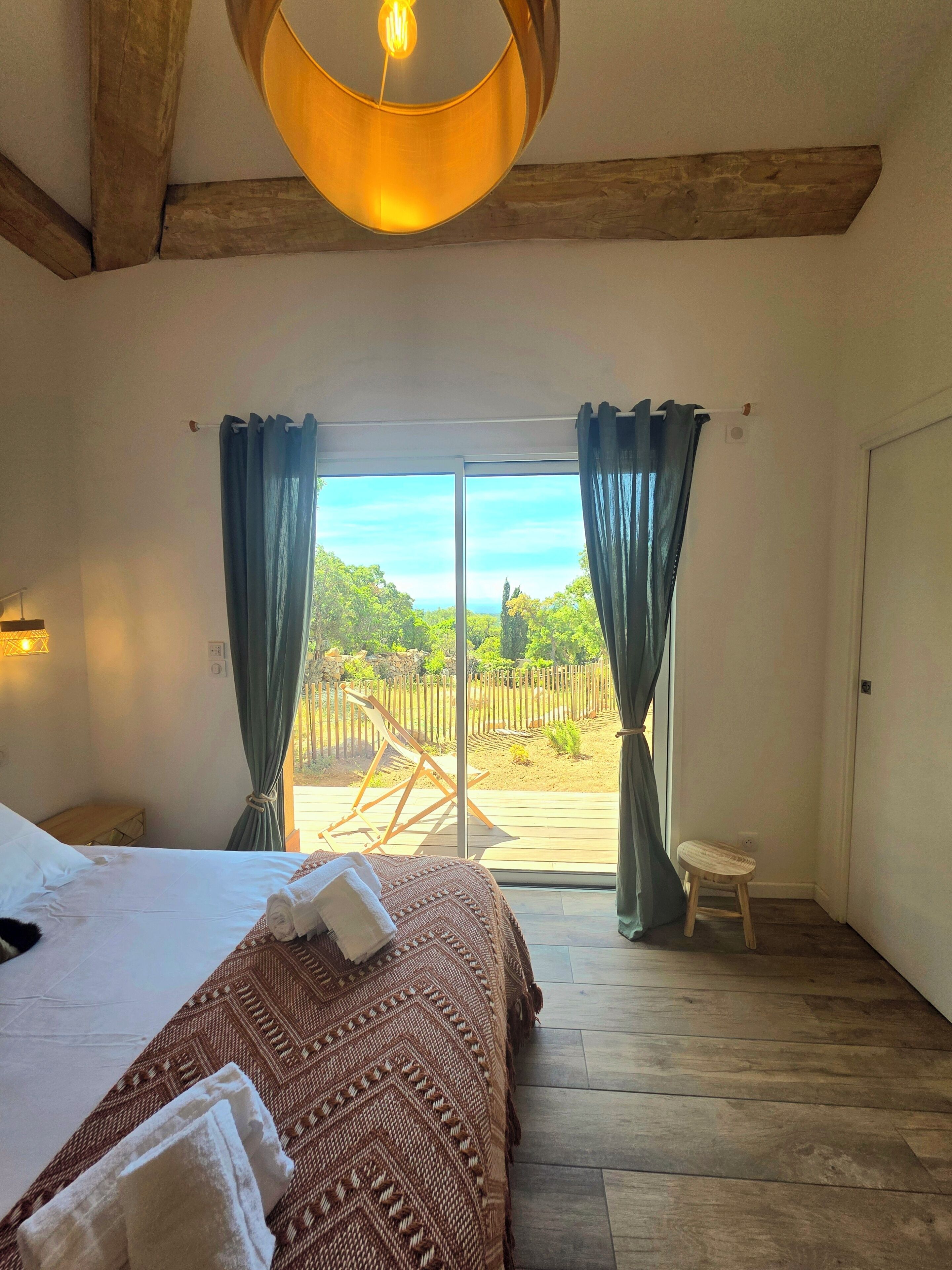 A bedroom with a bed, a wooden ceiling, a view through a glass door, and a small stool.