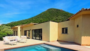 A swimming pool with lounge chairs, a building with large windows, and a hillside in the background. - Villa with pool, sea view, Corse-du-Sud (Monacia-d'Aullène)