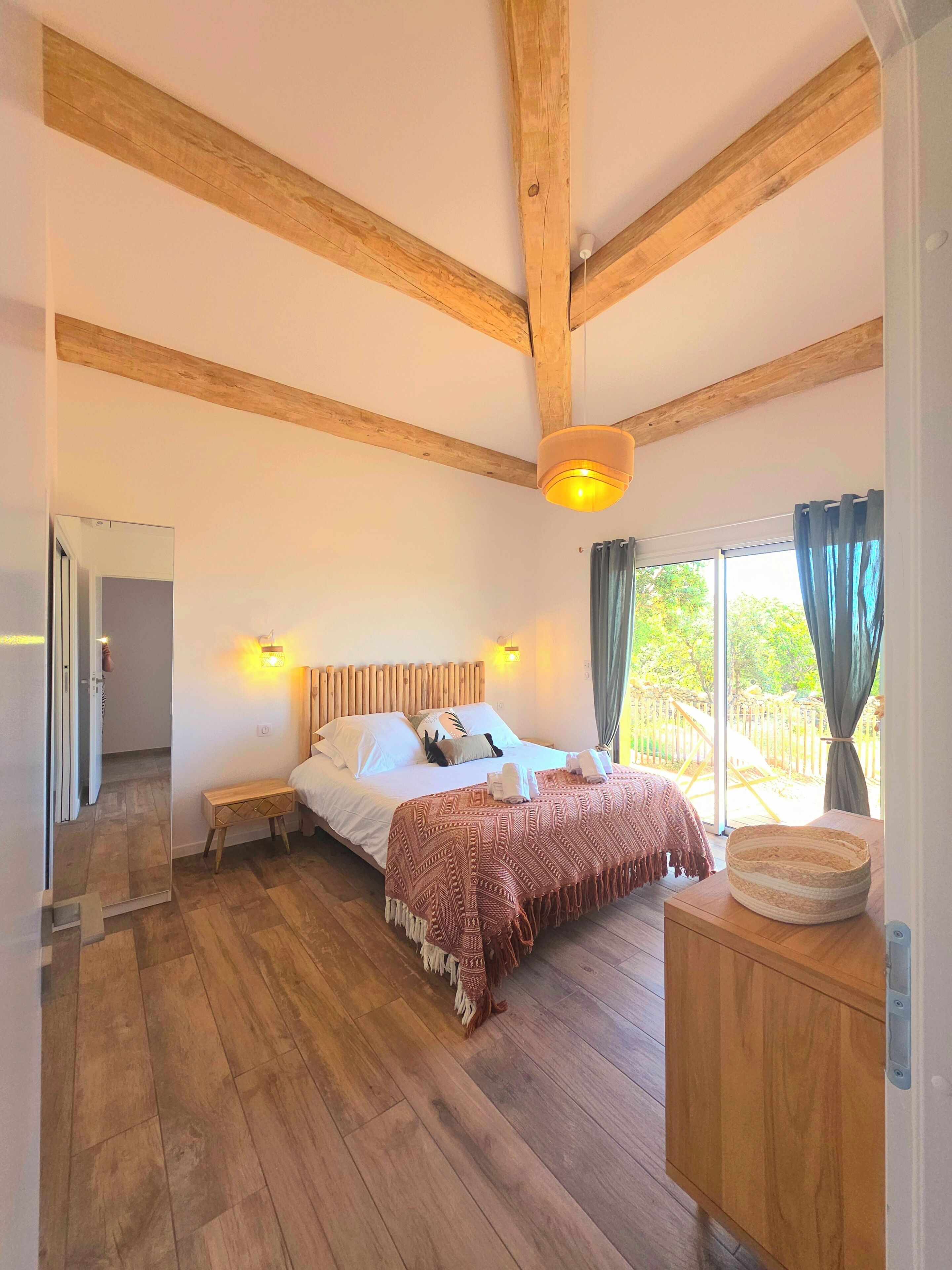 A spacious bedroom with a wooden ceiling, a large bed with a red blanket, a bedside table, and a view of the outdoors.
