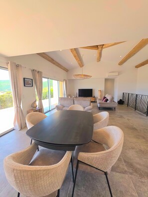 A spacious living room with a large black table, beige upholstered chairs, a sofa, and a TV.