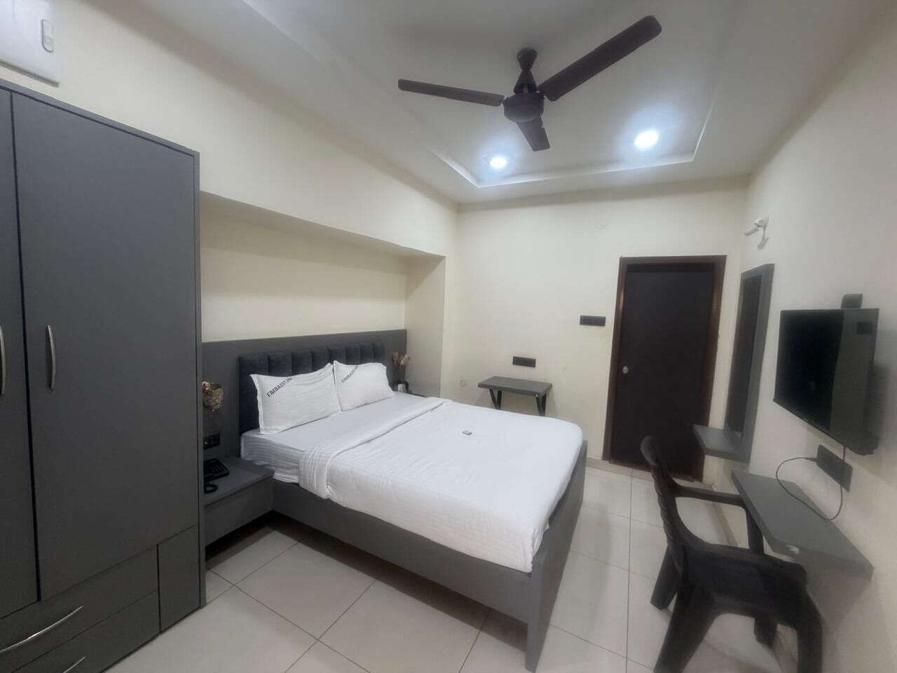 A bedroom with a bed, a desk, a chair, a wardrobe, a TV, and a ceiling fan.