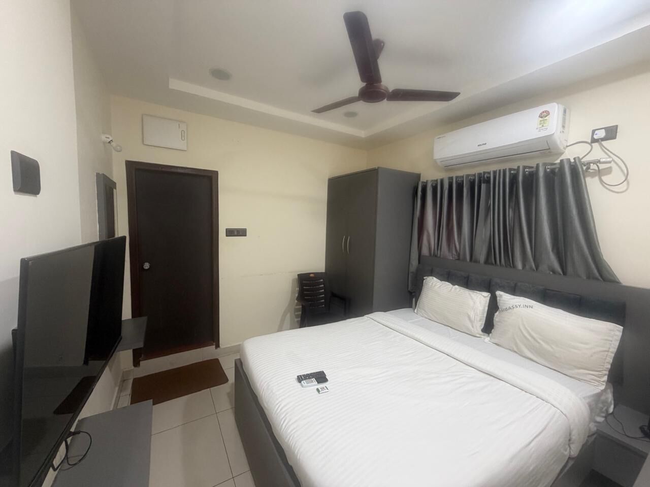 A hotel room with a bed, a desk, a wardrobe, a ceiling fan, an air conditioning unit, and a door.