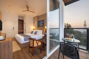 Habitación de hotel con cama, escritorio, silla, mesa con vasos y un jarrón, y balcón con vistas.