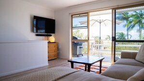 A living room with a sofa, a TV mounted on the wall, a wicker chest of drawers, and a sliding glass door leading to a balcony with a view of palm trees.