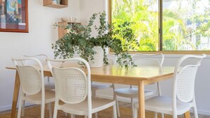 A dining area with a wooden table, white chairs, a woven rug, a potted plant, a framed picture, and a window with a view of greenery.