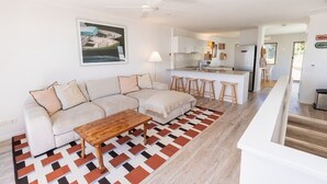 A modern living room with a sofa, a coffee table, a patterned rug, a kitchen area with stools, and a ceiling fan.