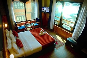 Quarto de hotel com cama, televisão, escrivaninha e vista para um edifício através de grandes janelas.