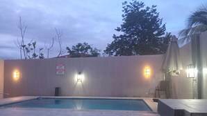 Una piscina con borde de piedra, dos luces empotradas en la pared, un cartel de advertencia y una palmera al fondo.