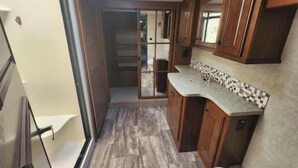 Un intérieur de camping-car compact comprenant un coin cuisine, des placards en bois et une porte coulissante donnant accès à un espace de rangement.