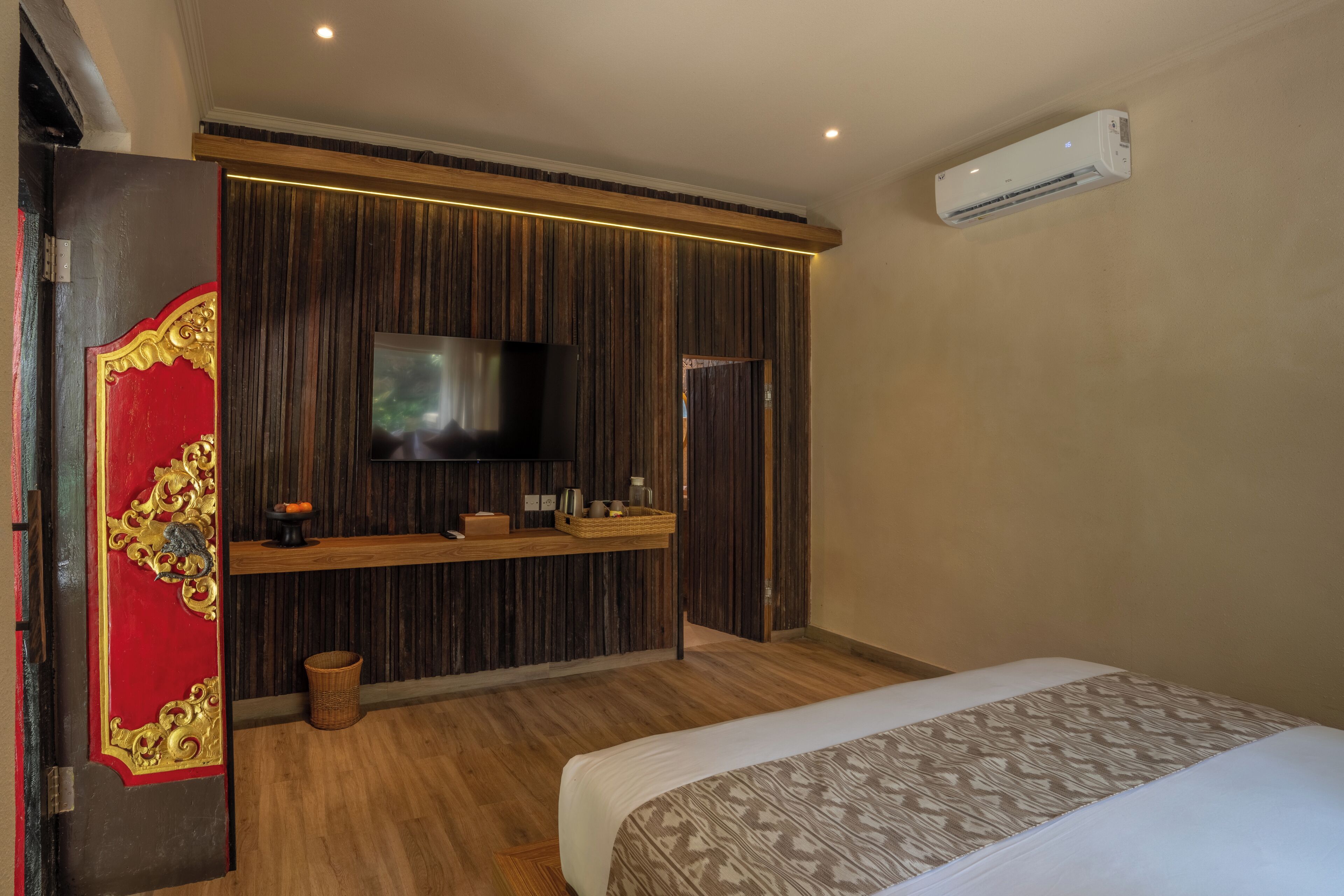 A hotel room with a bed, a TV, a wooden panel wall, a shelf with items, and an air conditioning unit.