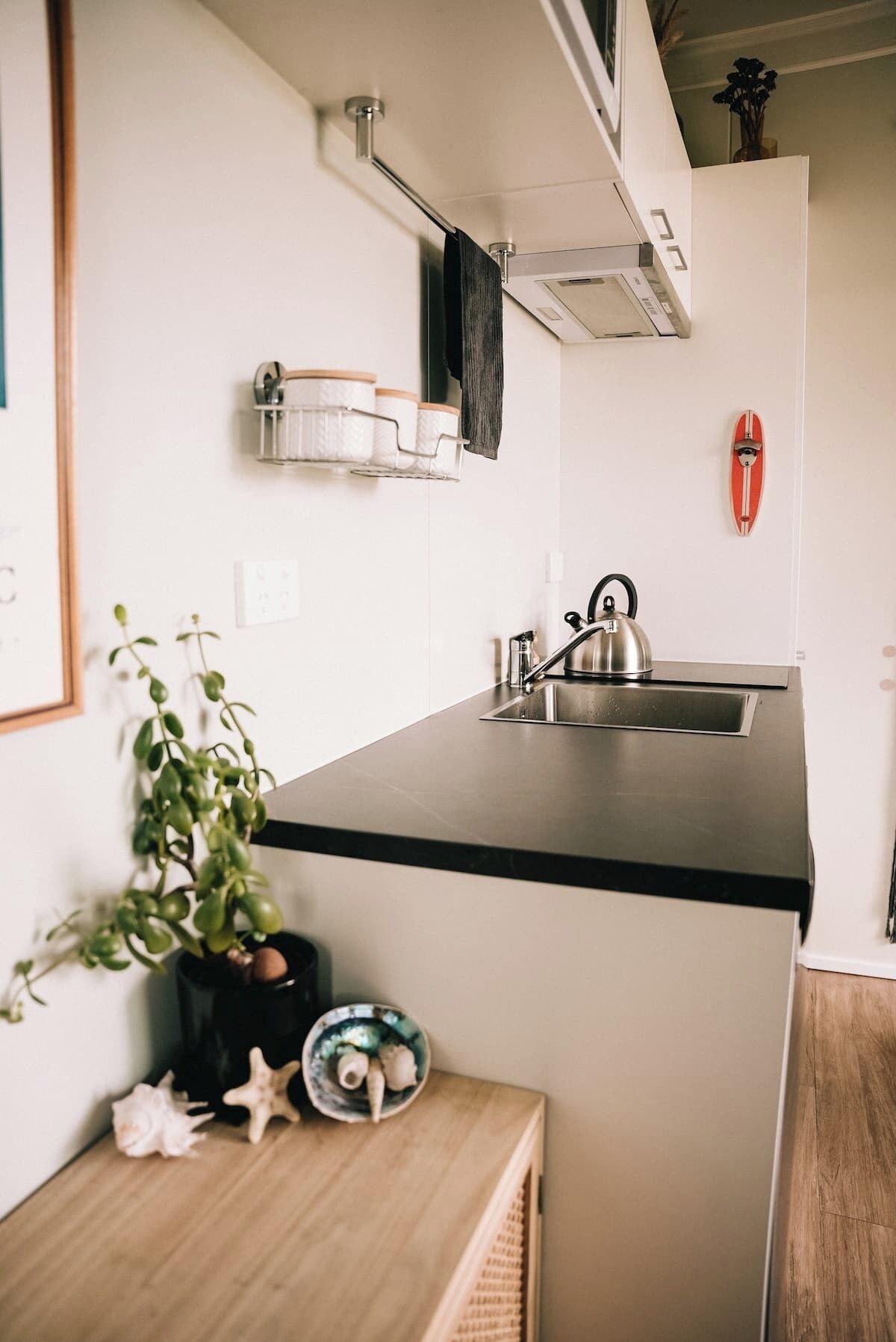 A compact kitchen with a black countertop, a kettle, a small sink, and a potted plant.