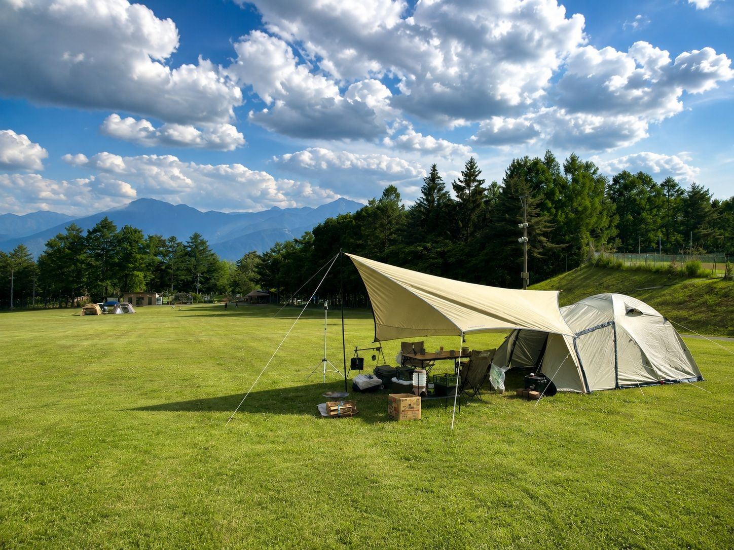 Un camping con tiendas de campaña, una mesa y sillas instalados en un campo cubierto de hierba, con montañas al fondo.