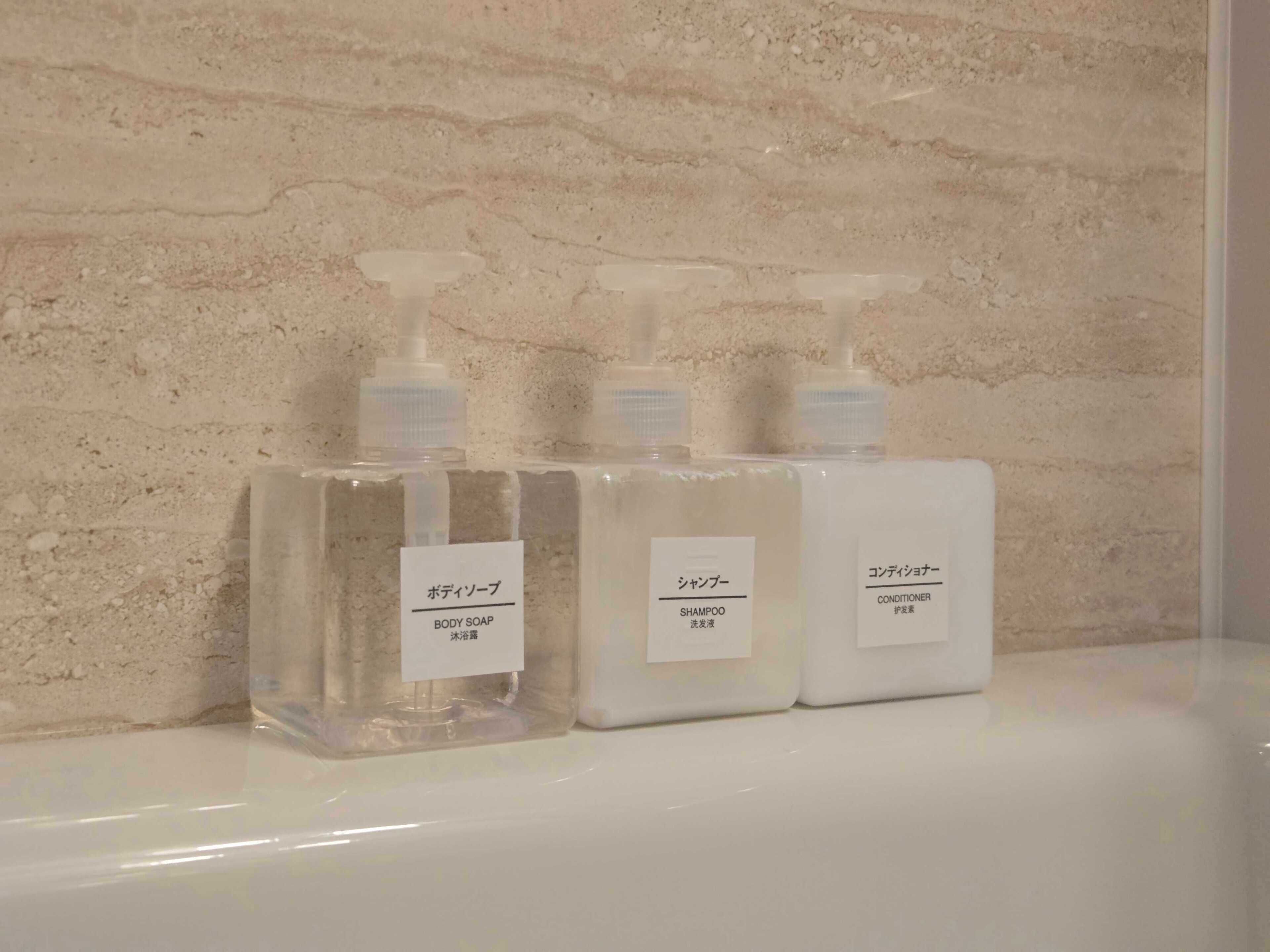 Three bottles of body soap, shampoo, and conditioner on a white surface against a textured wall.