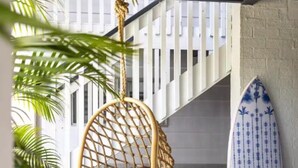 Een persoon zit in een hangstoel op een houten terras, met een surfplank tegen een muur geleund.