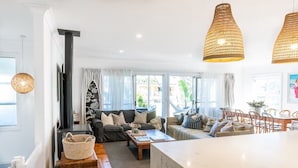 Een moderne woonkamer met een bank, salontafel en een eethoek. Er is een groot raam met gordijnen, een open haard en een hanglamp.