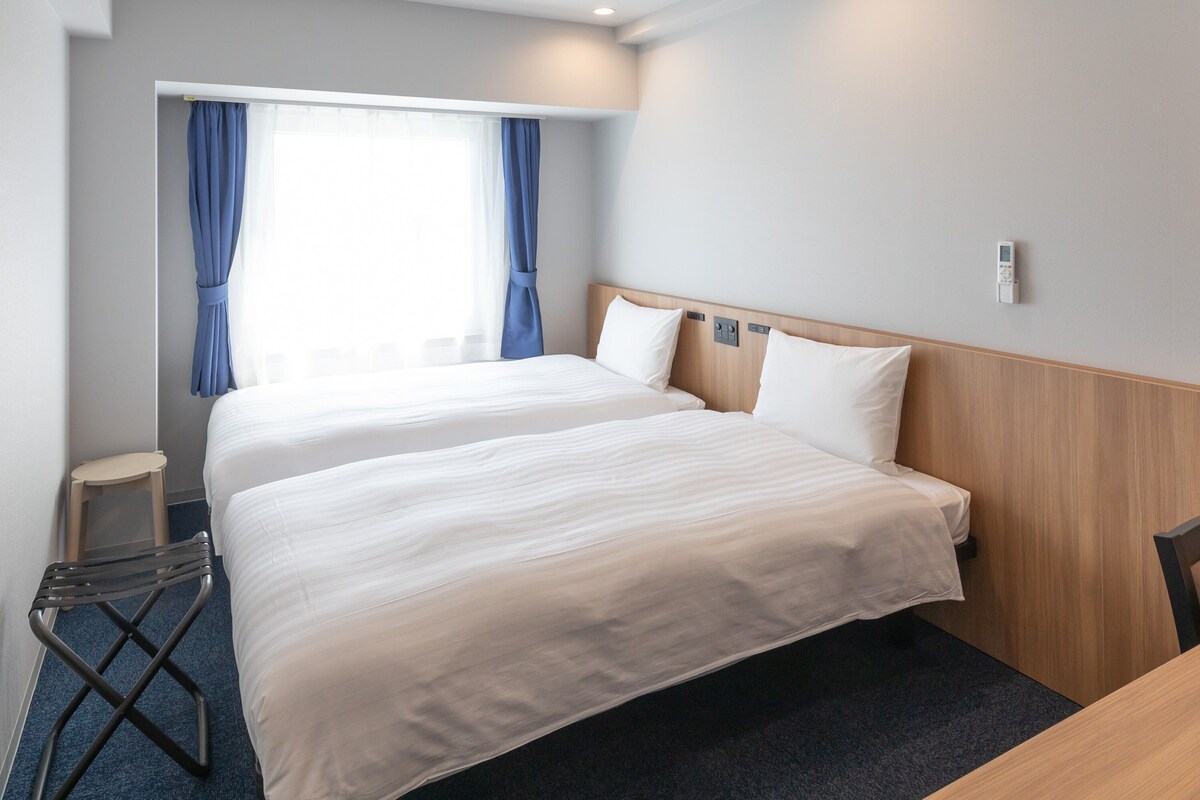 A hotel room with two beds, a small table, a chair, and a window with curtains.