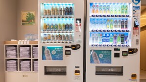 Dos máquinas expendedoras con diversas bebidas y aperitivos, un estante con toallas dobladas y un cuadro en la pared.