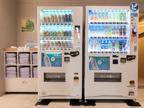Deux distributeurs automatiques proposant une variété de boissons et d’en-cas, une étagère avec des serviettes pliées et un tableau encadré au mur.