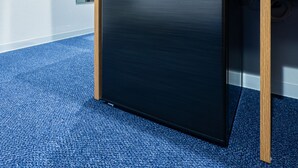 A compact, dark-colored refrigerator in a room with a blue carpet and wooden furniture.