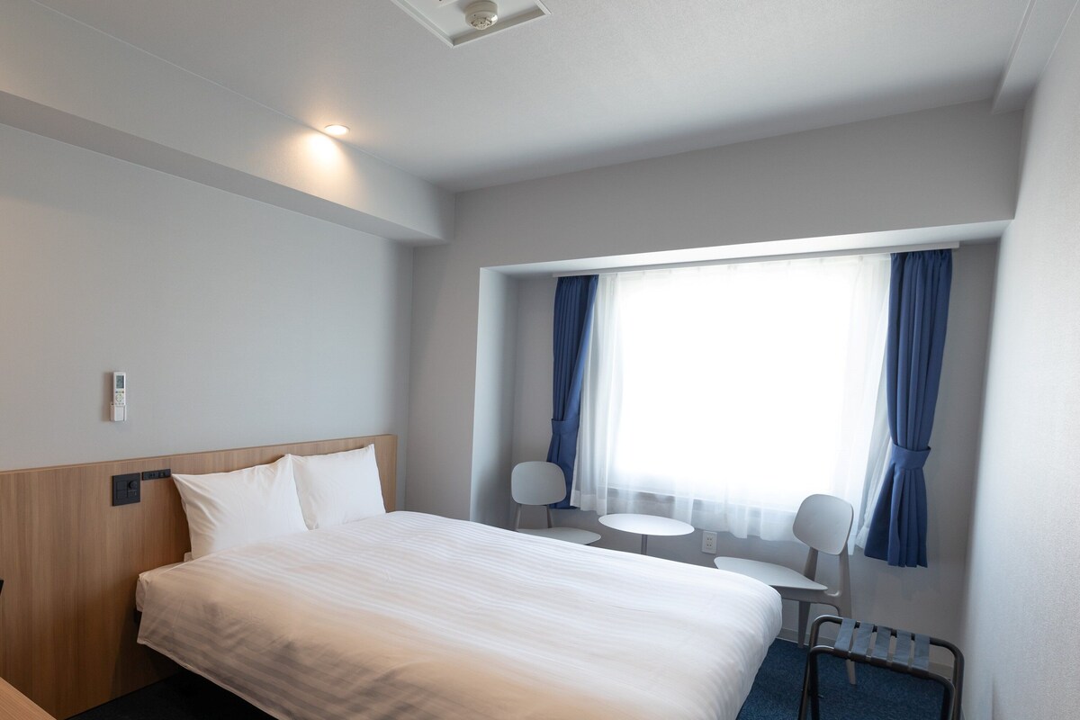 A hotel room with a bed, bedside table, lamp, and a window with curtains.