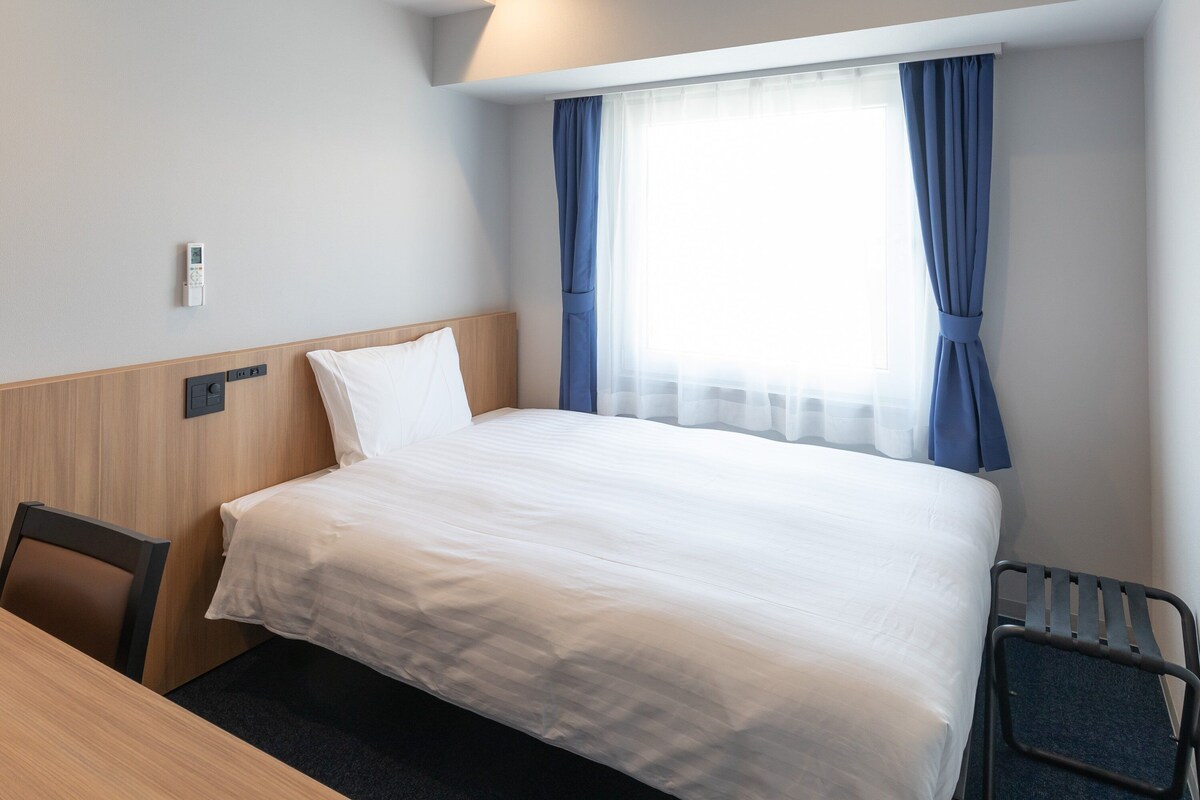 A hotel room with a bed, a desk, a chair, a window with curtains, and a thermostat on the wall.