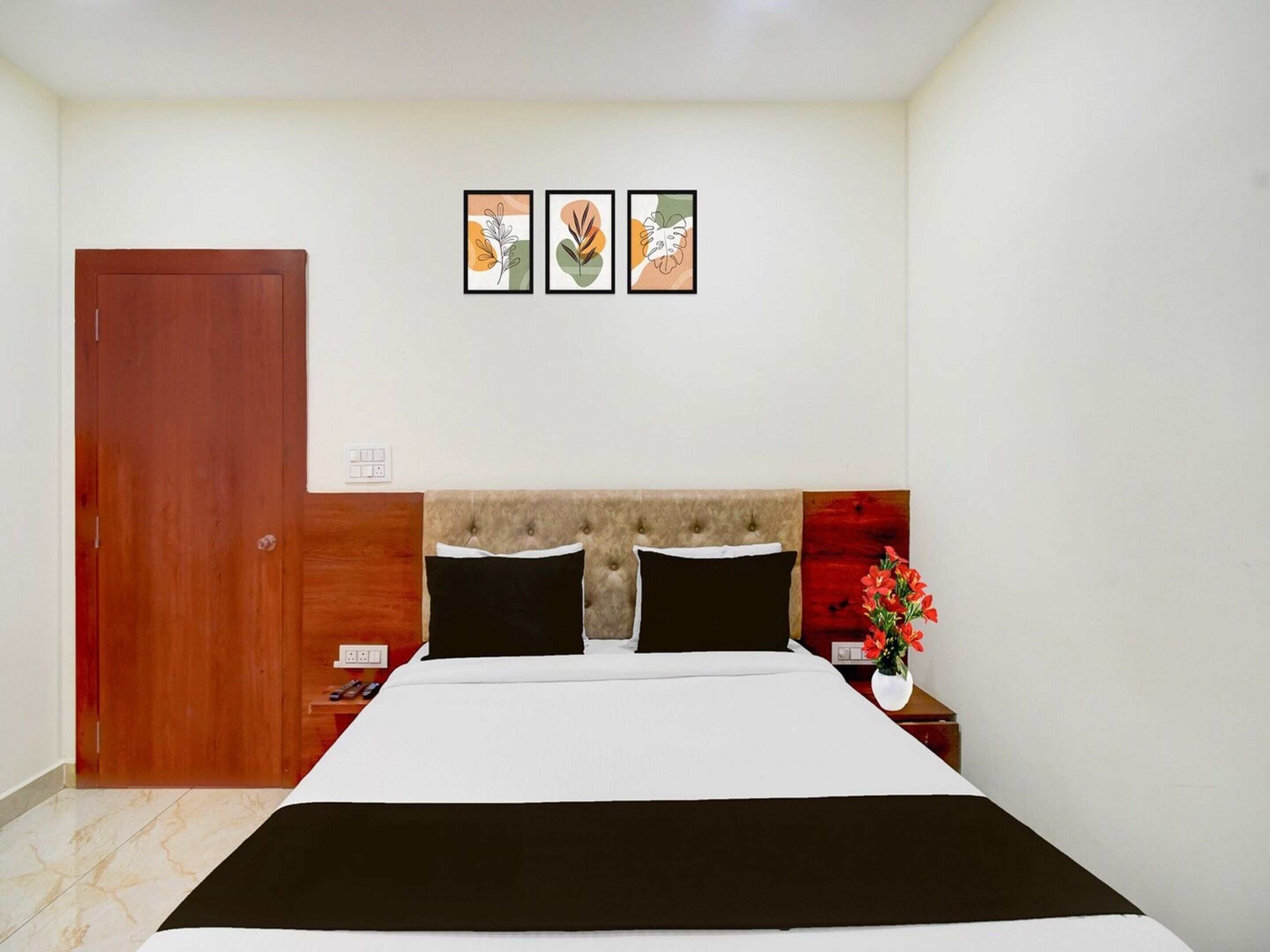 A bedroom with a bed, a wooden door, a nightstand, a vase with flowers, and three framed pictures on the wall.