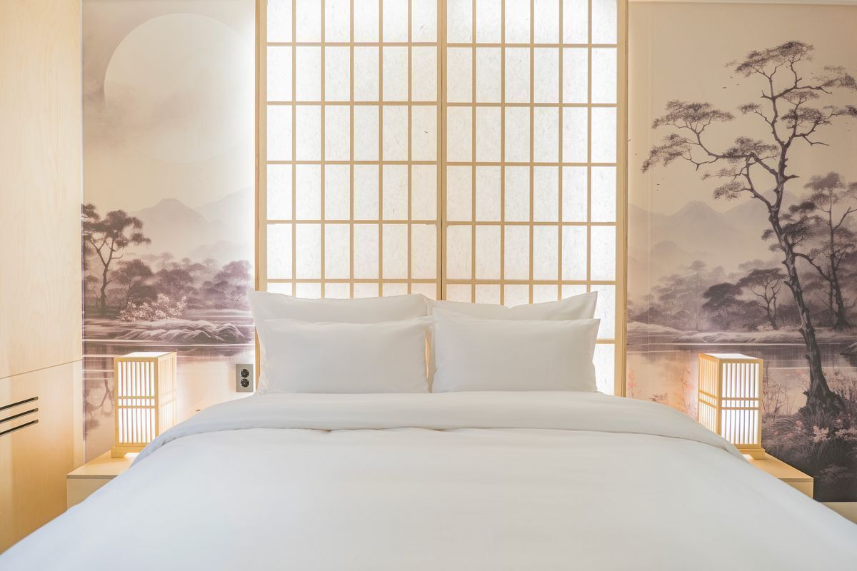 A bedroom with a bed, two bedside tables with lamps, a large window with a grid pattern, and a wall with a nature scene.