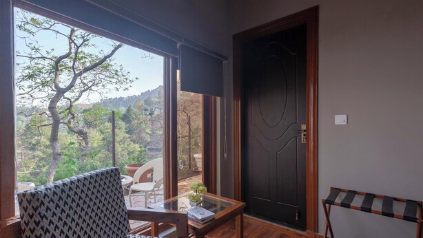 A room with a large window, a door, a chair, a small table, and a shelf. - Alaya Stays Amara Aether (Naina Range)