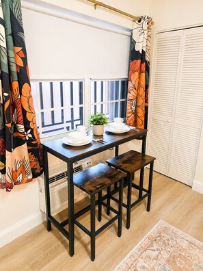 A small dining area with a table and two stools, a potted plant, and a window with blinds. - Cozy studio with Kitchen + Free Parking + Central location in Miami. (Miami)