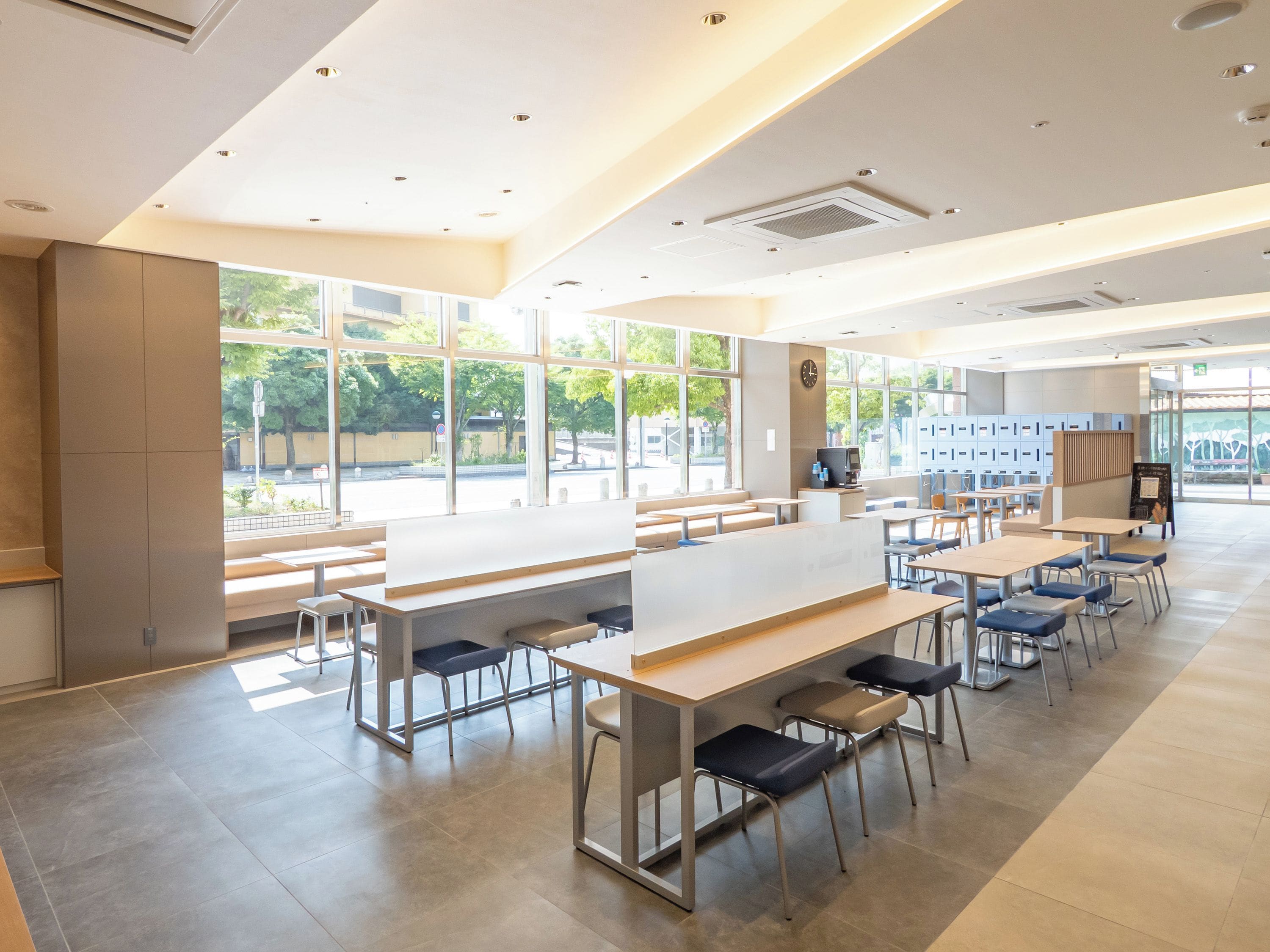 Una cafetería moderna con mesas y bancos largos, ventanas amplias y un reloj en la pared.