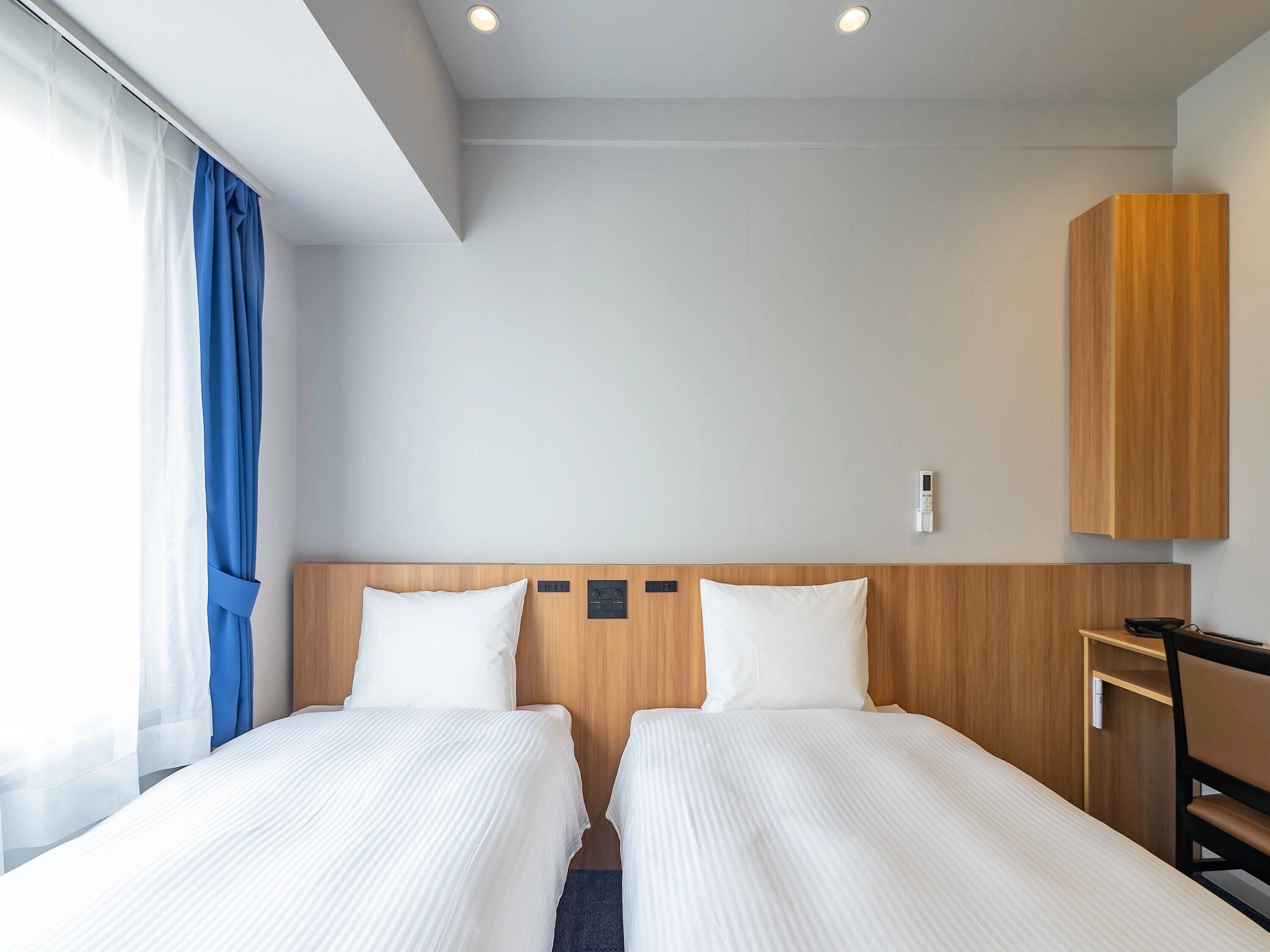 A hotel room with two beds, a wooden headboard, a desk with a chair, and a window with curtains.