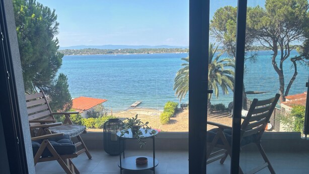 Un balcón con vista al mar, sillas de madera, una pequeña mesa con una planta y suelo de baldosas.