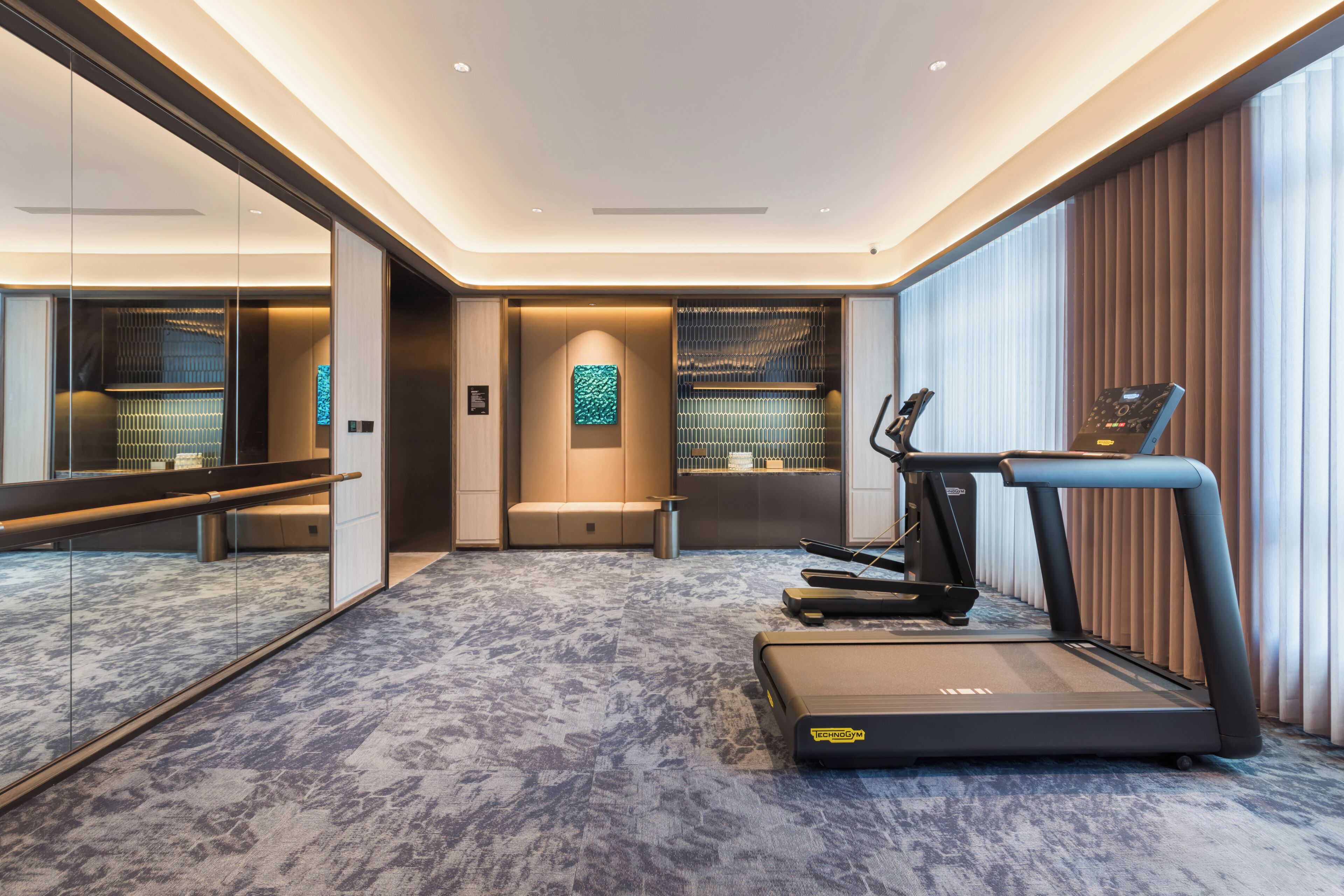 Ein modernes Hotelzimmer mit Laufband, einer Sitzecke und einem großen Spiegel, der den Raum wiederspiegelt.
