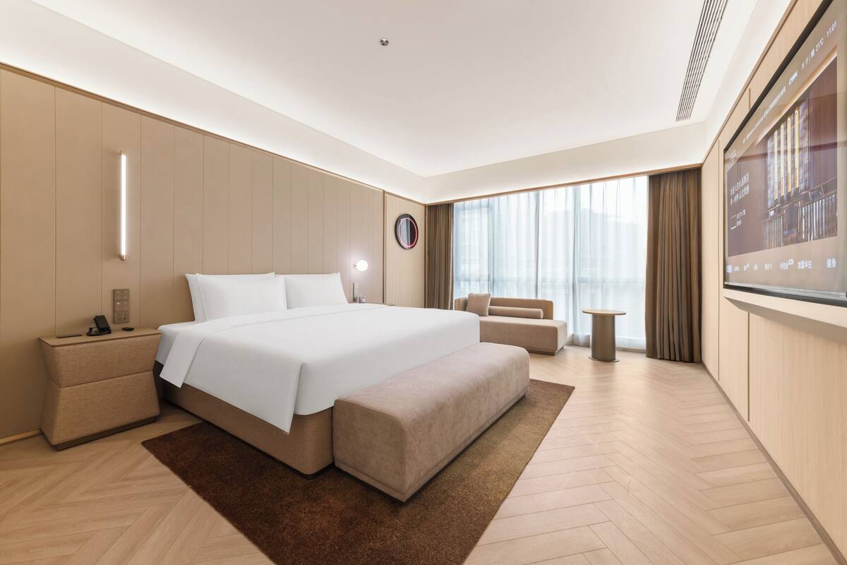 A modern hotel room with a large bed, a bedside table, a small sofa, a round table, and a large window with curtains.